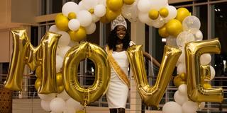 76th miss central state university raven golliday stands in her royal court gear and crown in an arch of gold and white balloons while behind big balloons spelling the word move