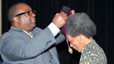 an african american man in a grey suit places a lanyard and medallion over the head of an african american woman in a leopard print shirt