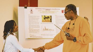 A student researcher shakes hands with an alumni attendee of Scholarly Research Activities Day at Central State University. The title of the research project shown on a posterboard in the background is, "Viral Prevalence of Small Carpenter Bees Among Different Farm Landscapes"