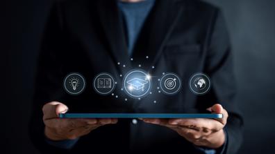 a person wearing a black suite jacket and blue undershirt holding a laptop with icons of a lightbulb, book, graduation hat, bullseye, and earth above it