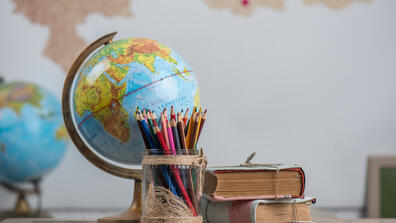 A world globe, colored pencils in a glass jar, and two stacked well used books placed on a desk with a second globe and wall map in the background.