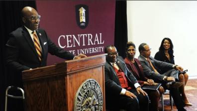 Morakinyo A.O. Kuti, Ph.D., speaks at a podium during a ceremony announcing him as the 10th president of central state university in wilberforce ohio as other leaders look on in excitement