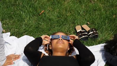 a central state university student wearing eclipse glasses while laying on a blanket in the grass at the seed to bloom botanical and community garden