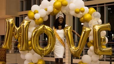 76th miss central state university raven golliday stands in her royal court gear and crown in an arch of gold and white balloons while behind big balloons spelling the word move