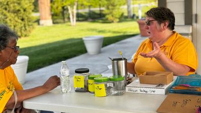 two people in gold shirts talk at a table with jars of seeds