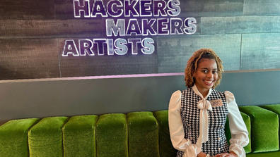 a young african american woman wearing professional dress sitting on a green couch with a sign saying hcakers makers artists on the wall