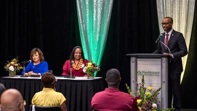 dr. arletha mcswain and another person sit at a table while a man stands at a podium nearby