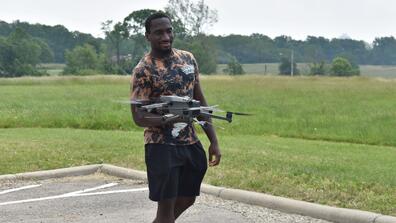 a young black man wearing a camo shirt with a drone flying in front of him in a parking lot beside a field