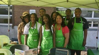 eight youth and adults wearing green aprons at a farmers market