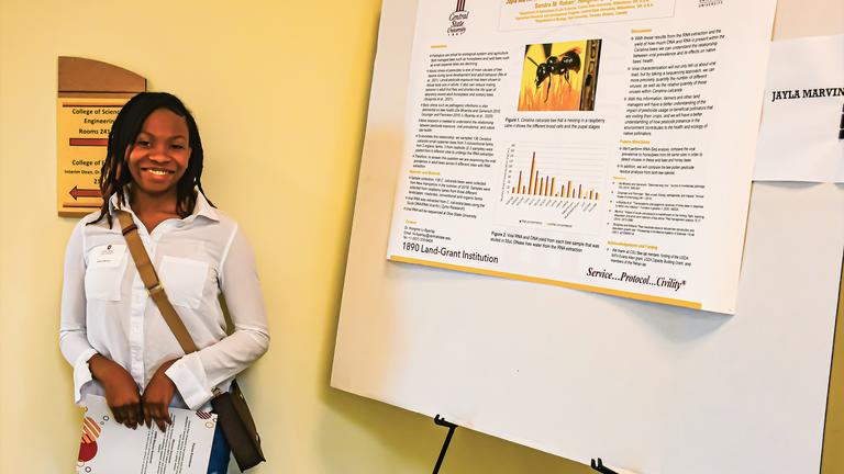 a Black person stands beside a presentation board with the results of their research at Central State University