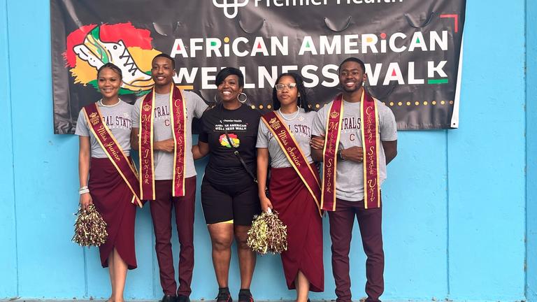 four Central State University Royal Court members stand in front of a Premier Health sign for the African American Wellness Walk, pictured with the event organizer