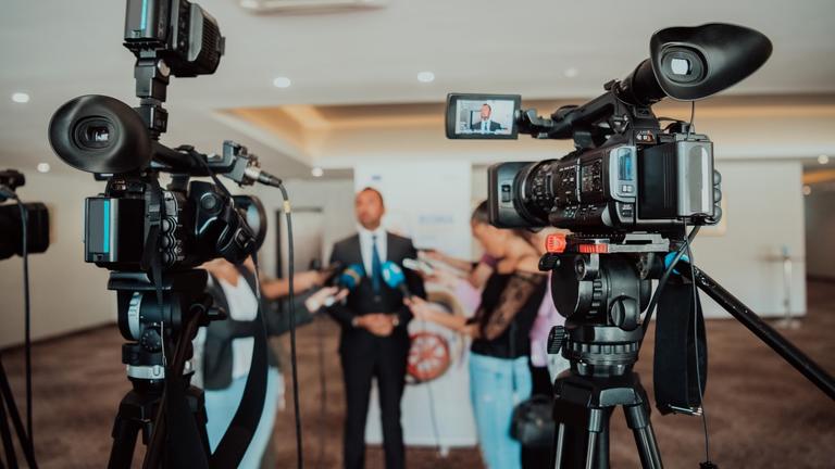 several people conduct on-camera interviews while standing around the subject being interviewed with two cameras in the foreground