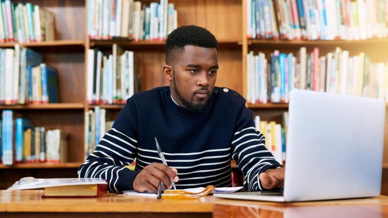 an African American student studies in a library using books and a laptop