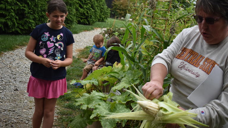 a child walks toward Clare Thorn, extension associate, holding an ear of corn
