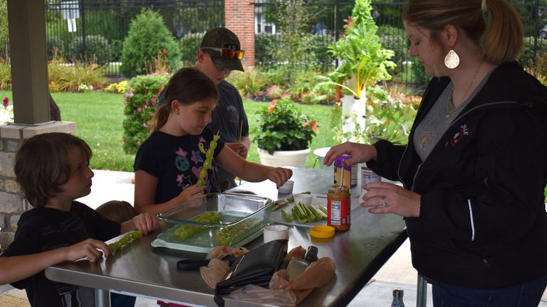 children stand at a table while an adult helps them get a snack of grapes, celery, and peanut butter