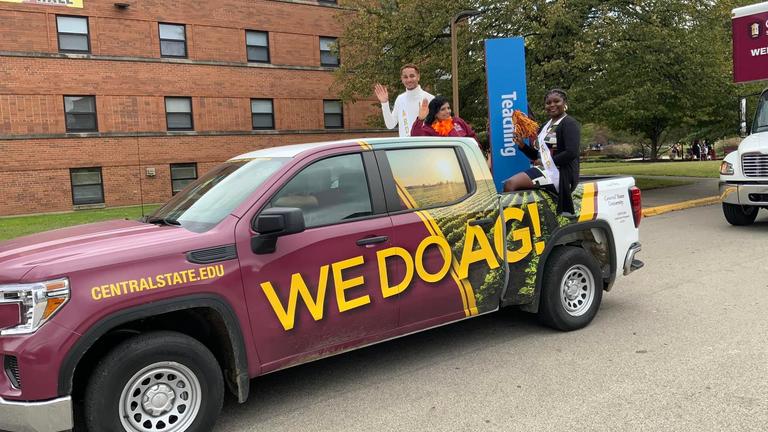 people wave during a parade while riding in the back of a truck with the words we do ag!