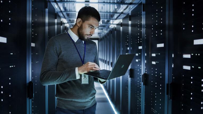 IT Technician Works on a Laptop in Big Data Center full of Rack Servers. He Runs Diagnostics and Maintenance, Sets up System