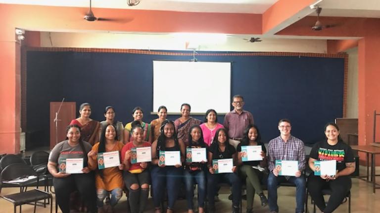 A student group poses in front of the stage of a meeting room in India.