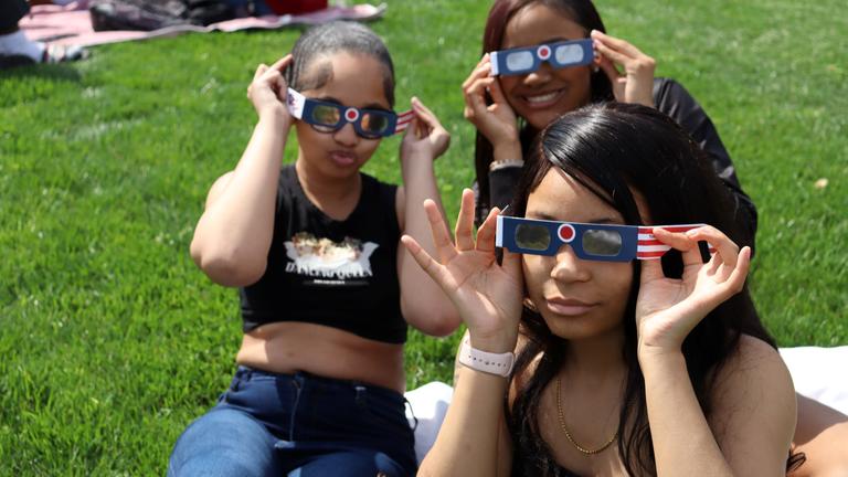 three young women wearing eclipse glasses while sitting on the grass
