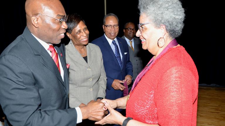 a man in a suit holds the hands of a woman in reddish orange while two people smile at them in the background