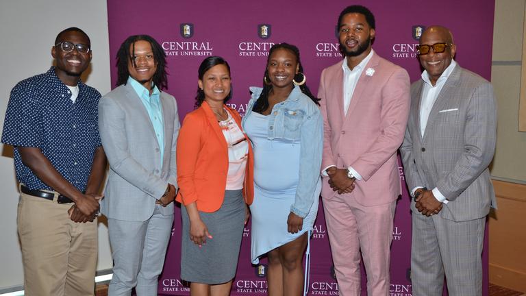 six people in professional dress stand in front of a central state university step and repeat