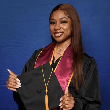 A Black woman, Asiah Robinson, stands before a blue wall wearing graduate regalia