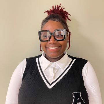 Christian Jackson, a student at Central State University, smiles. Christian is wearing a white shirt with a vest and large glasses and has braided hair pulled back.