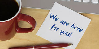 Image of a coffee cup next to a keyboard, ink pen and postie note with the words "We are here for you!"