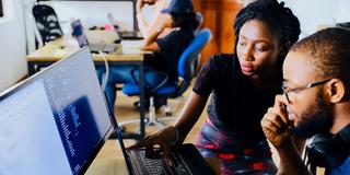 Two Black college students work on a computer with more students studying in the background