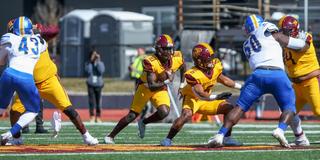 Football players on a field. One team is wearing gold uniforms with maroon highlights and the other is wearing white jerseys with blue game pants