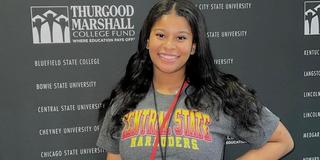 A young Black woman with long Black hair wearing a Central State University T-shirt in front of a backdrop for the Thurgood Marshall College Fund Yardi program
