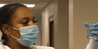 A young Black environmental engineer, Jasmine Walker, wearing a white lab coat and blue protective gloves holding a test tube for water quality testing