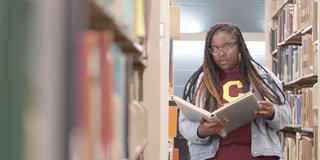 a student wearing a Central State University t-shirt and denim pants and jacket reads a book while browsing the stacks at the Hallie Q. Brown Memorial Library