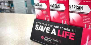 three narcan kits in a pharmacy. a sign in front of them says everyone has the power to save a life. complete your first aid kit. easy to use.