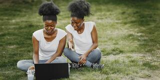 African American twin women sit in a grassy area with a laptop