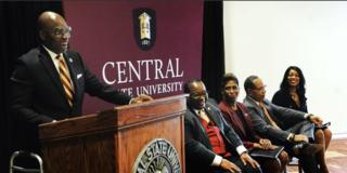 Morakinyo A.O. Kuti, Ph.D., speaks at a podium during a ceremony announcing him as the 10th president of central state university in wilberforce ohio as other leaders look on in excitement