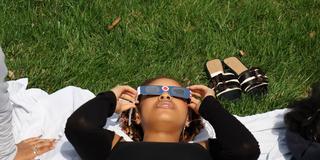 a central state university student wearing eclipse glasses while laying on a blanket in the grass at the seed to bloom botanical and community garden