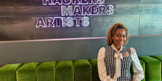 a young african american woman wearing professional dress sitting on a green couch with a sign saying hcakers makers artists on the wall