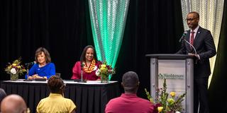 dr. arletha mcswain and another person sit at a table while a man stands at a podium nearby