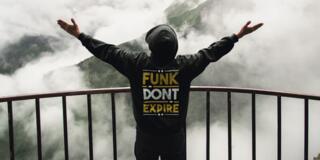 a person in a funk don't expire hoodie stands with arms spread in front of a mountain