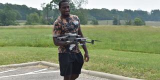 a young black man wearing a camo shirt with a drone flying in front of him in a parking lot beside a field