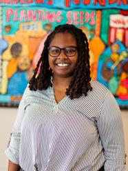 A young Black woman with dark black hair smiles at the camera with a brightly colored mural behind her