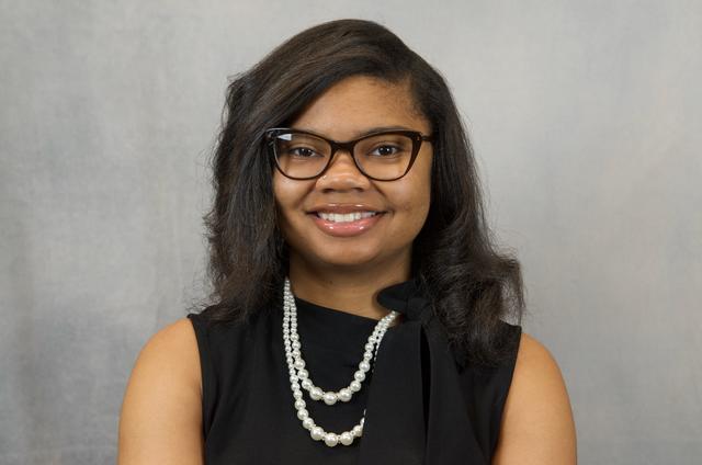 A young Black woman in black with white pearls and glasses smiles at the camera