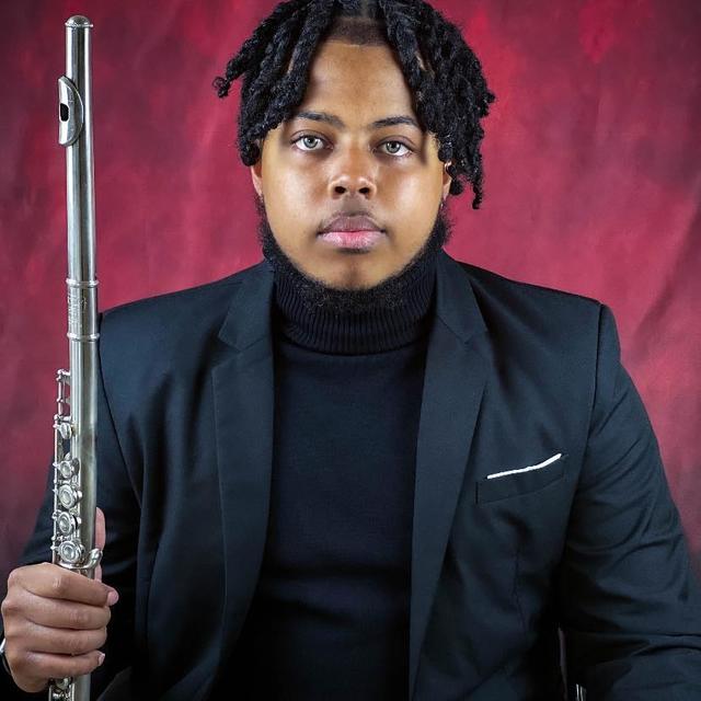 A young African American man wearing black holds a flute vertically with a maroon background