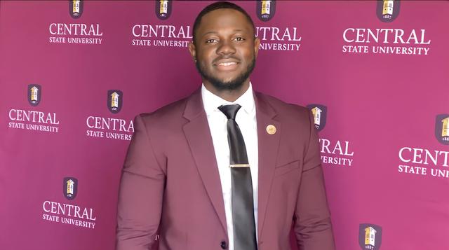 A young Black man in a maroon suit and black tie smiles in front of a Central State University backdrop
