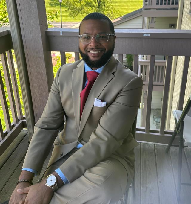 Chad Tillman sits in a suit and tie on a porch during a sunny day