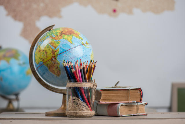 A world globe, colored pencils in a glass jar, and two stacked well used books placed on a desk with a second globe and wall map in the background.