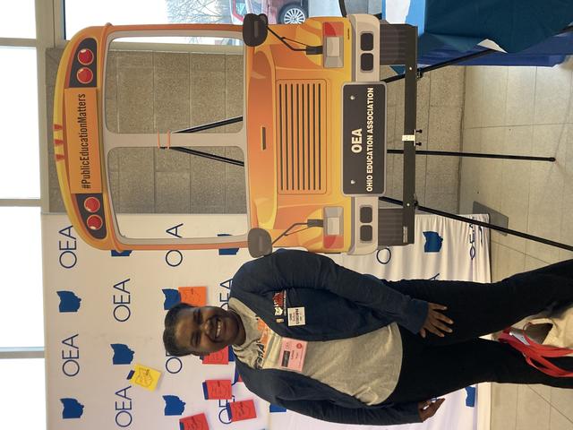 a young Black woman stands beside a cutout poster of a school bus with the ohio education association logo on a step and repeat in the background