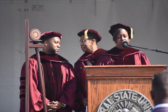 natalia harris speaks into microphone with christopher wyche and jacqueline gamblin smile at each other behind her on stage at commencement