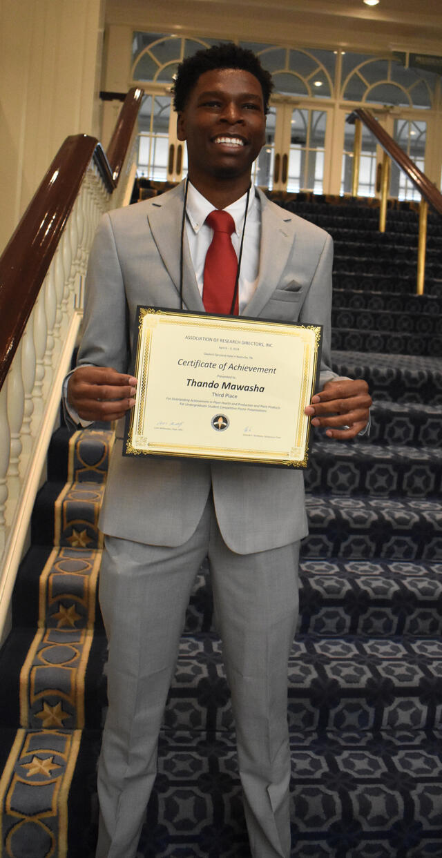 Thando Mawasha holding a certificate and wearing a grey suit and red tie smiling broadly on stairs